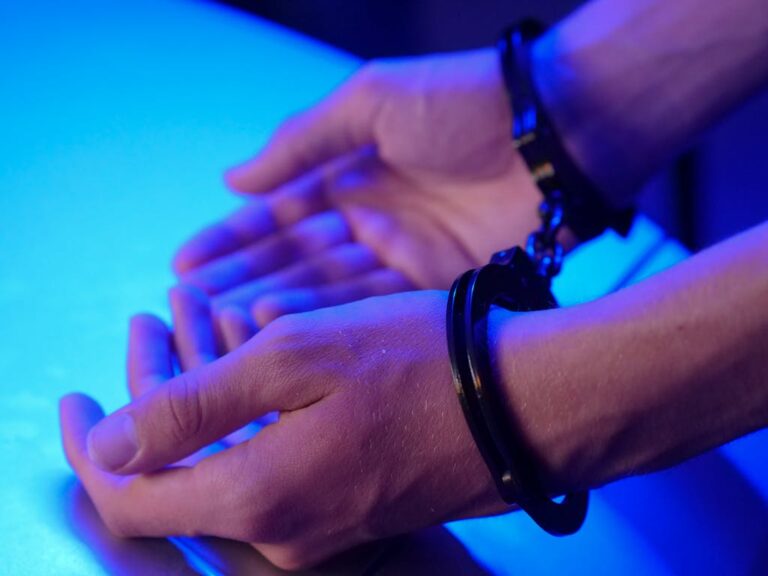 Close-up image of hands in handcuffs resting on a table under blue lighting depicting law enforcement